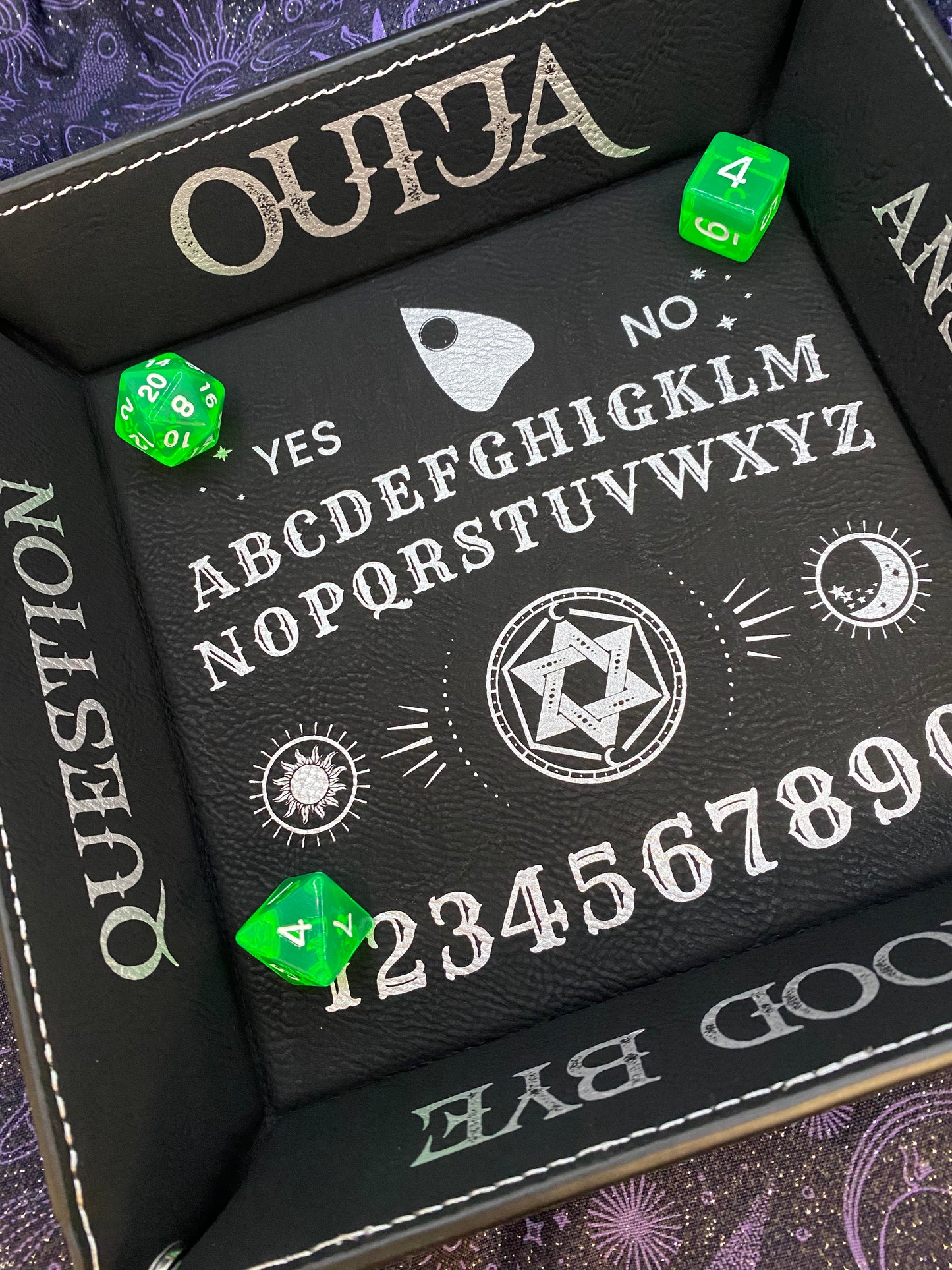 Black leather dice tray with silver laser-engraved Ouija board design, featuring alphabet, numbers, sun and moon symbols, and a planchette graphic. Green dice displayed inside the tray.