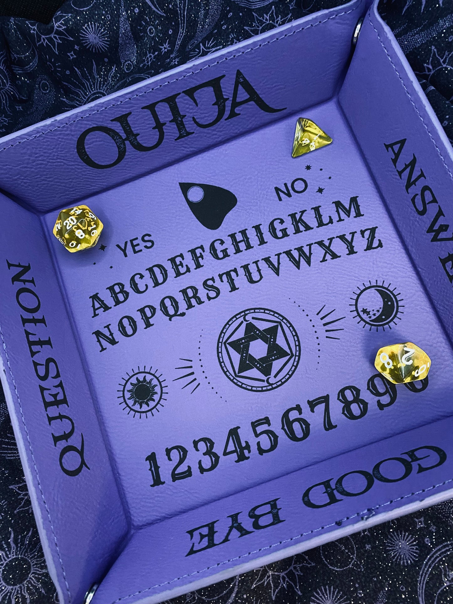 purple leather dice tray with black laser-engraved Ouija board design, featuring alphabet, numbers, sun and moon symbols, and a planchette graphic. Gold dice displayed inside the tray.